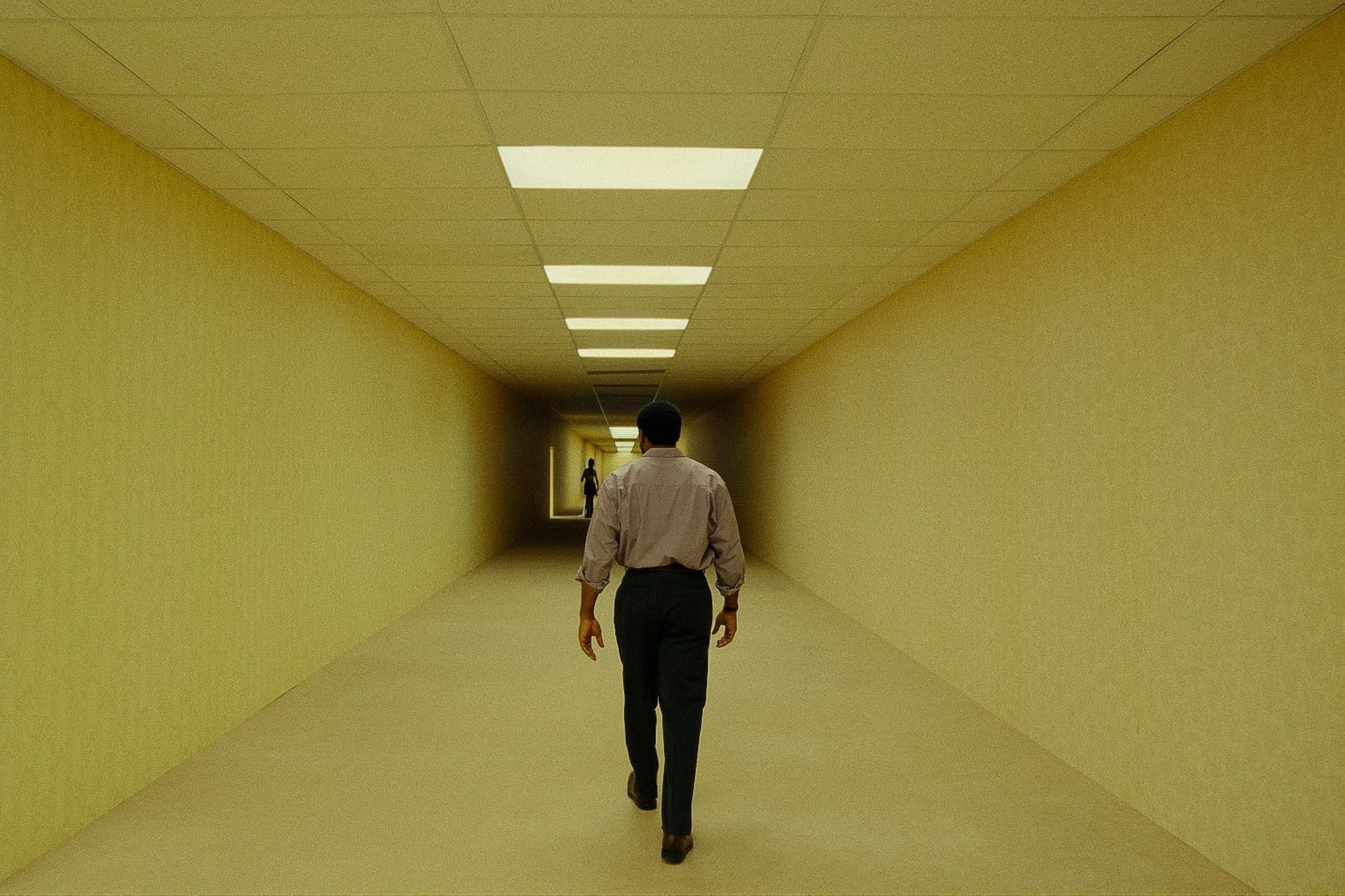 Un hombre camina por un pasillo amarillo iluminado con luces fluorescentes en Backrooms